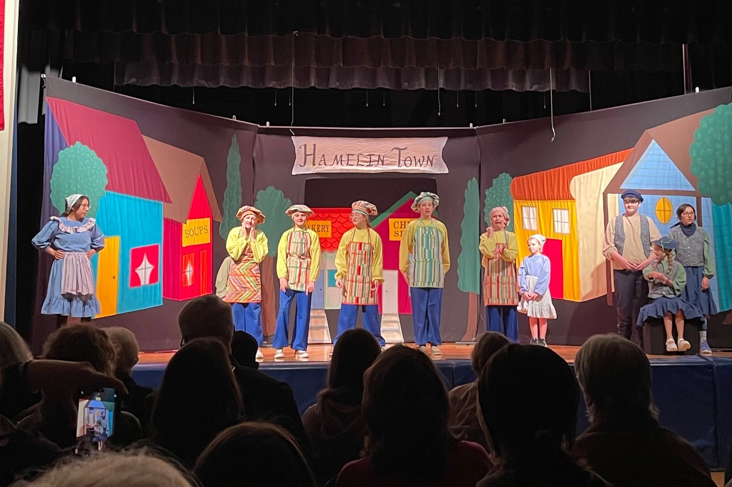 Children performing a play on stage with a backdrop of a colorful town scene, including buildings labeled 'Soups,' 'Bakery,' and 'Chimney Sweeps,' and a sign reading 'Hameen Town.'