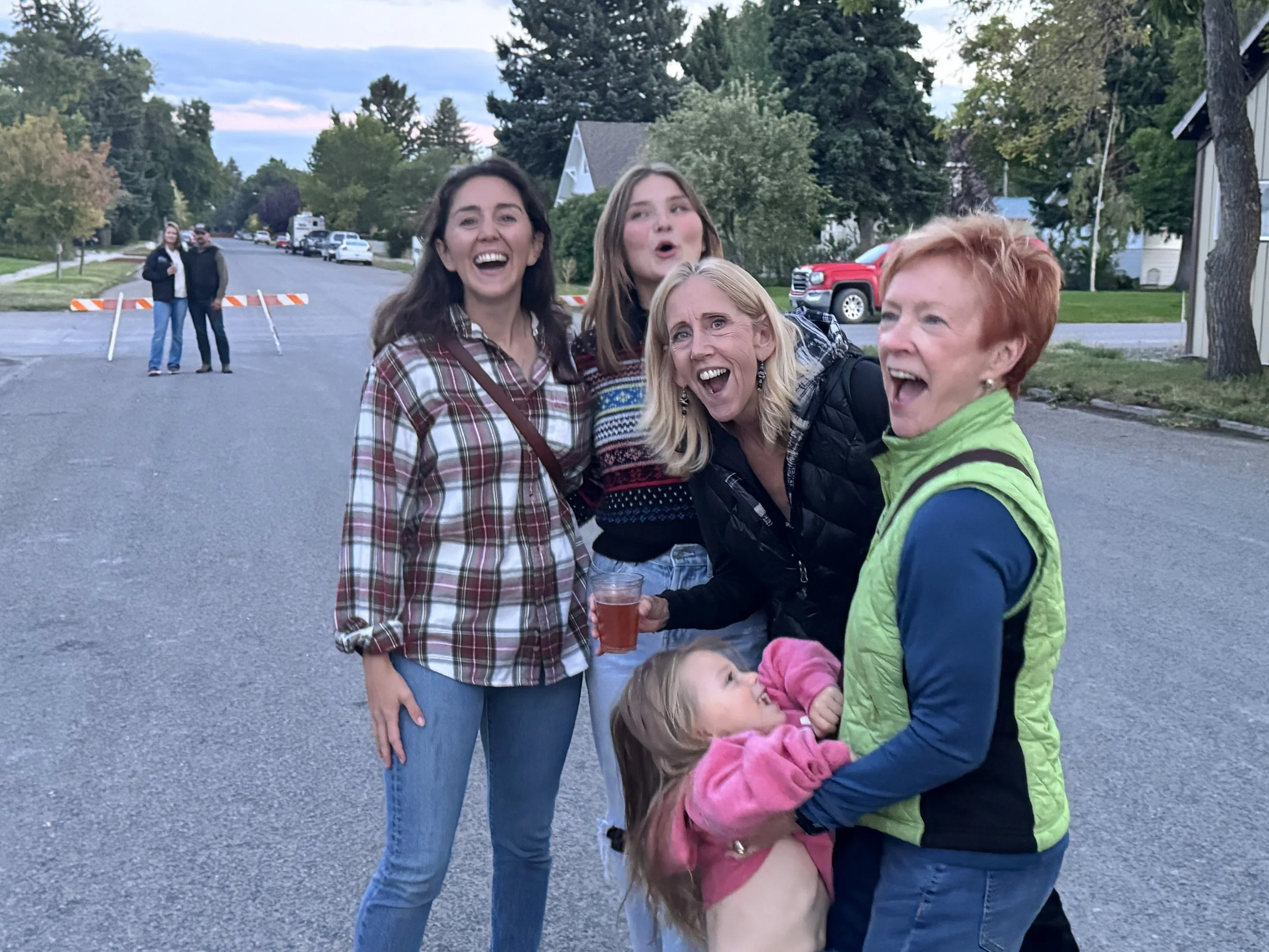 Group of five women and a young girl laughing and smiling outdoors on a street during the evening, with some houses, trees, and parked cars in the background.