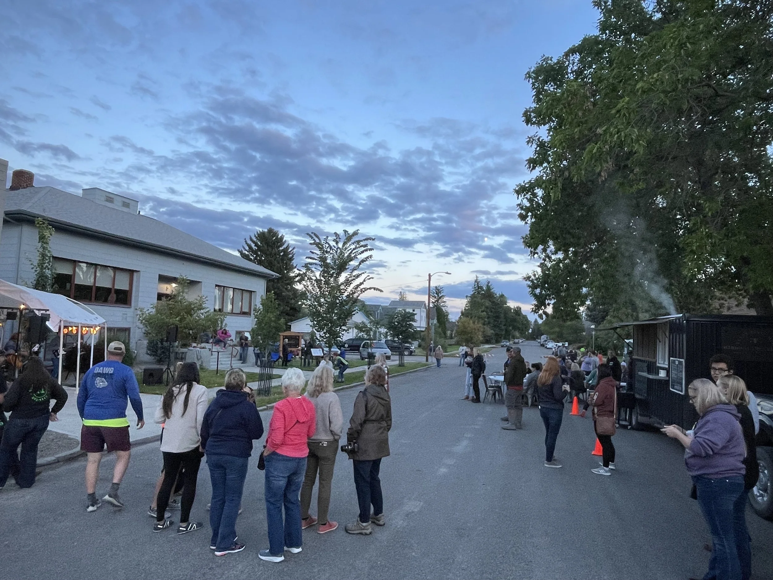 A street scene during an outdoor evening event, with people gathered around and talking. There is a food truck on the right with steam rising from it and a small stage on the left with musicians performing under a large tent.