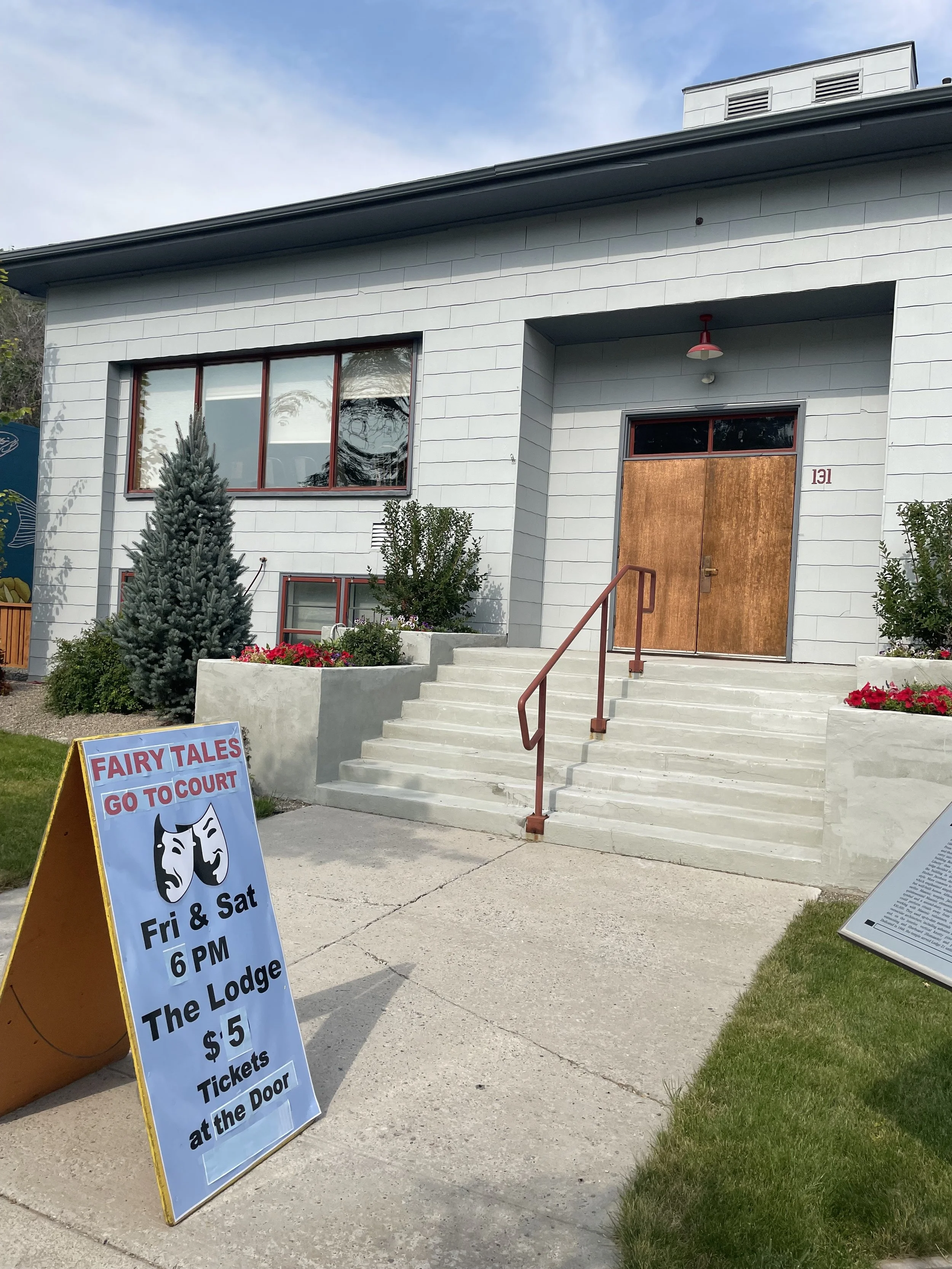 Sign advertising fairy tales performance at The Lodge on Friday and Saturday at 6 PM, tickets $5 at the door, placed on a sidewalk in front of a modern building with stairs, bushes, and flowers.