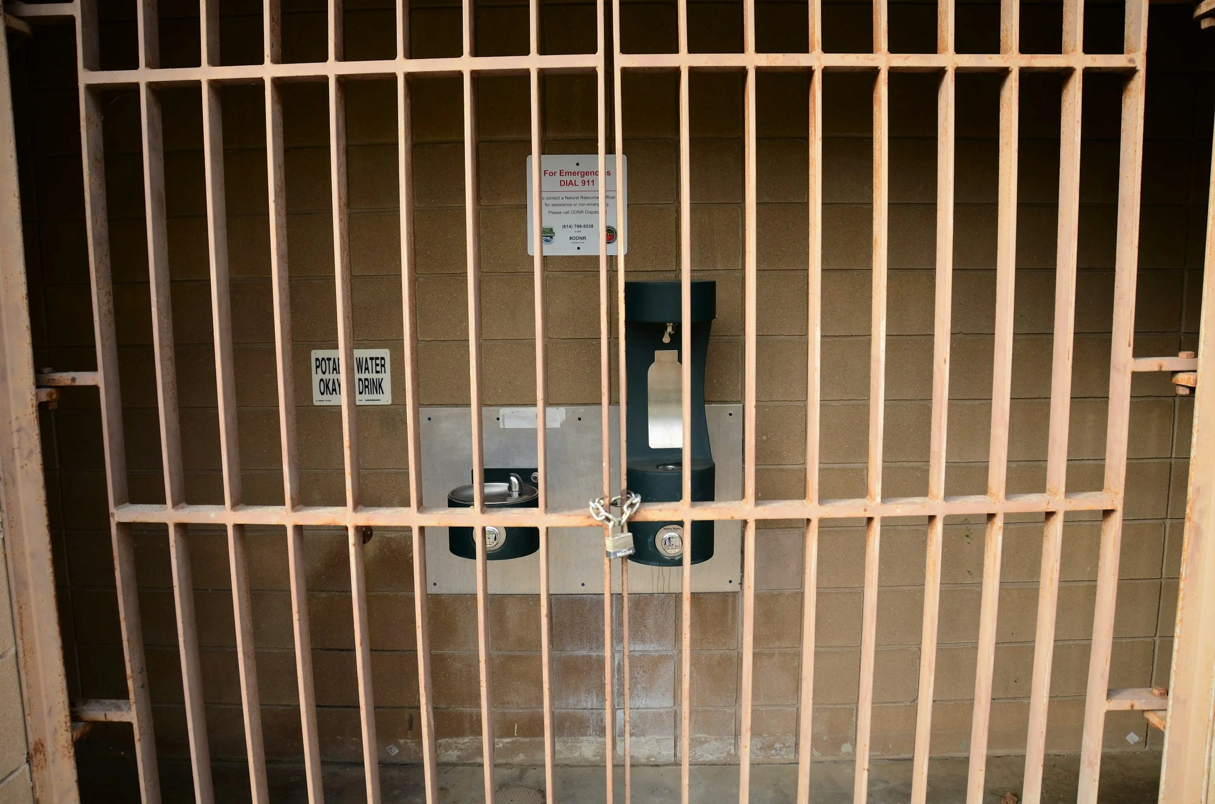 Public water fountain behind metal bars in a secured area, with signs indicating water and potability, and a lock on the bars.