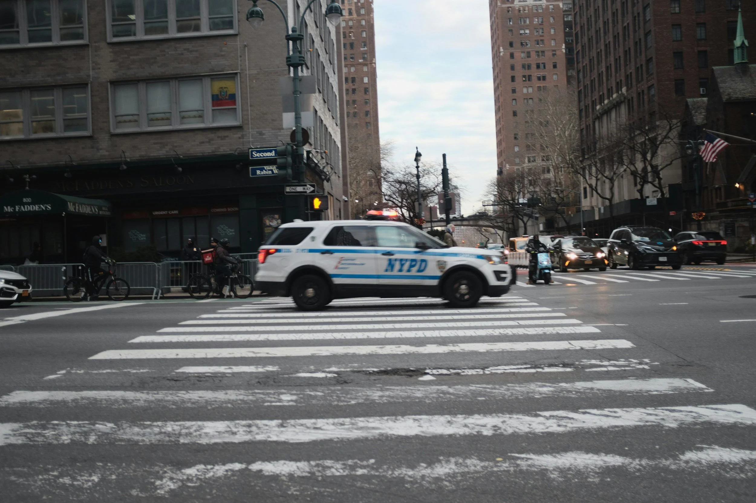 A city street with a white NYPD police SUV in the middle of a crosswalk. Several cyclists are waiting at the crosswalk, and there are multiple cars on the street. Tall buildings and a cloudy sky are visible in the background.