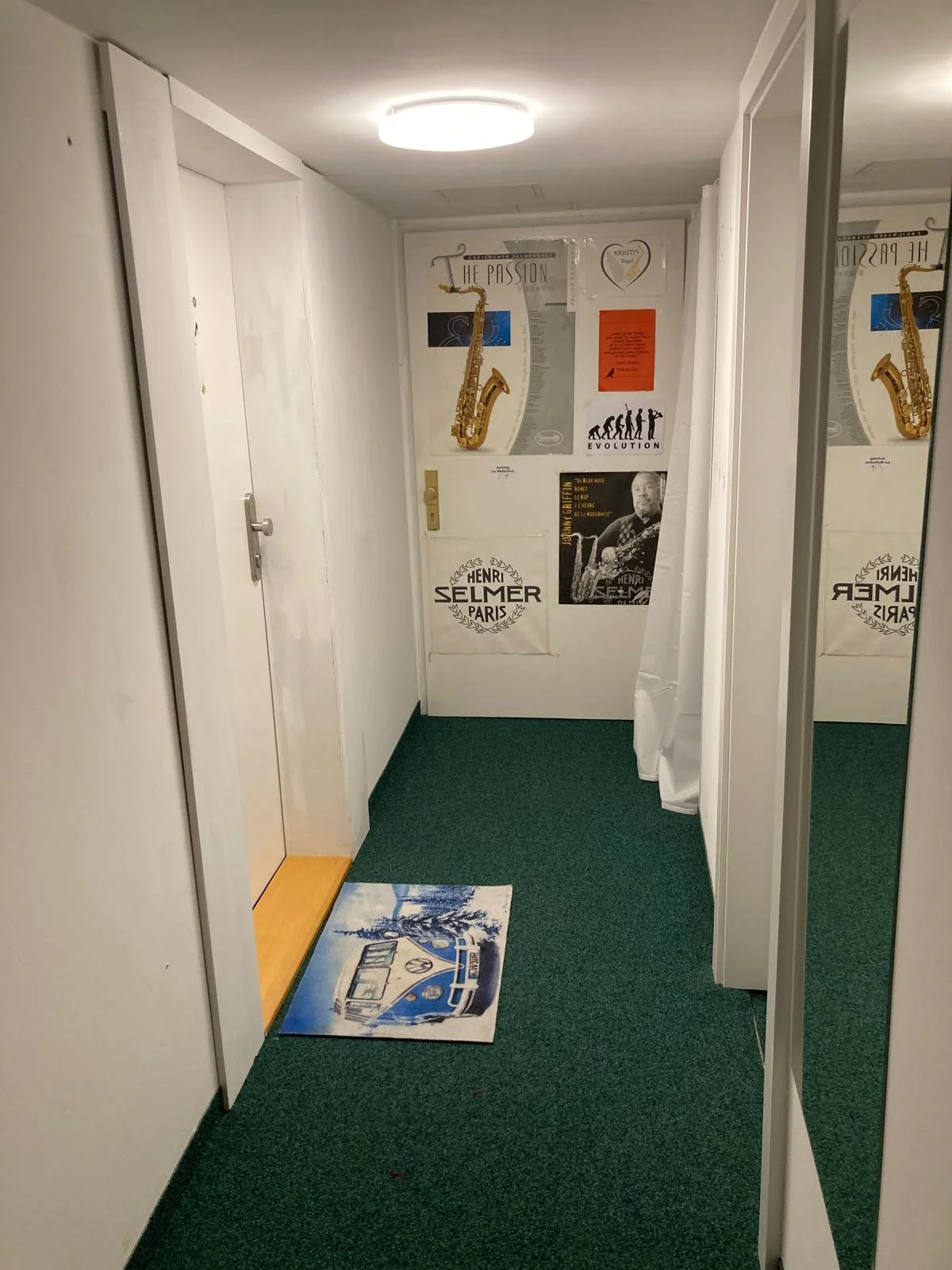 Flur mit grüner Teppich, weiße Wände, Poster, eine Tür, ein Teppich mit VW Bus Grafik und ein Spiegel an der rechten Wand.