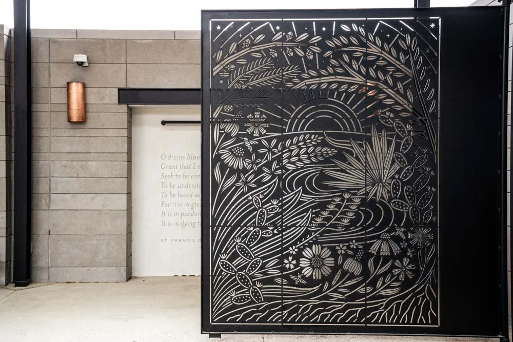 Decorative metal entry gate with floral and nature-themed cutout design, partially obscuring a wall with a quote on a white surface.