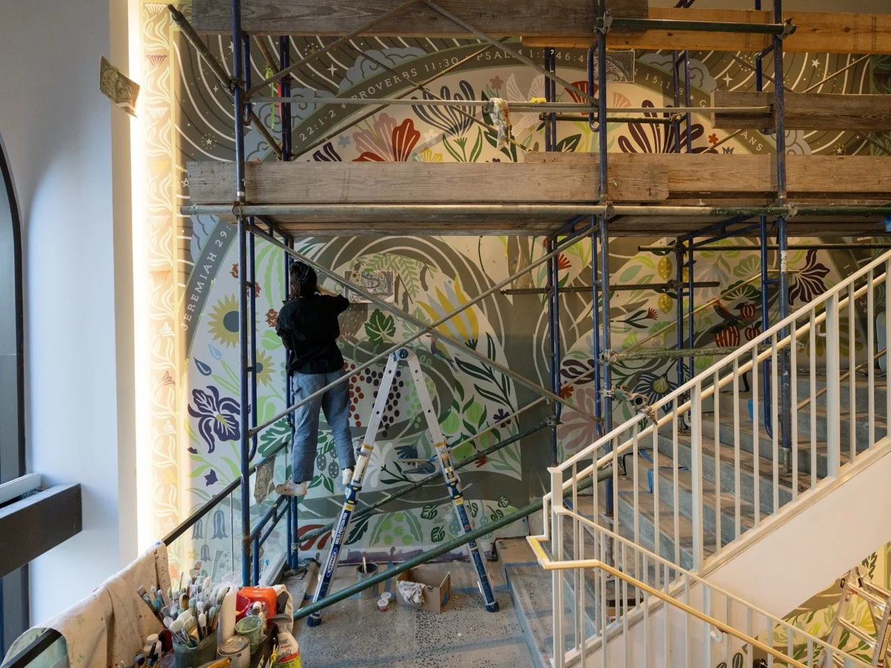 A person working on a mural or wall art installation on a large colorful floral and geometric patterned wall, with scaffolding and a ladder in an indoor space.