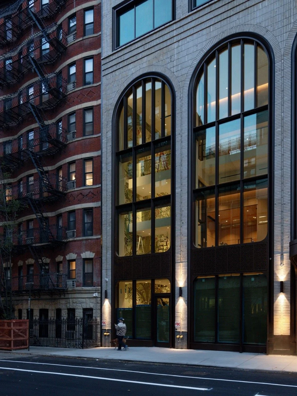 Modern multi-story building with large arched glass windows and exterior lighting at dusk, located next to an older red brick building with fire escapes.
