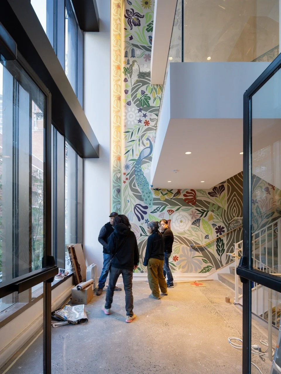 Interior of a building with large windows, a colorful floral and peacock mural on the wall, and a staircase. A group of five people are standing near the mural, possibly discussing or working on a project.