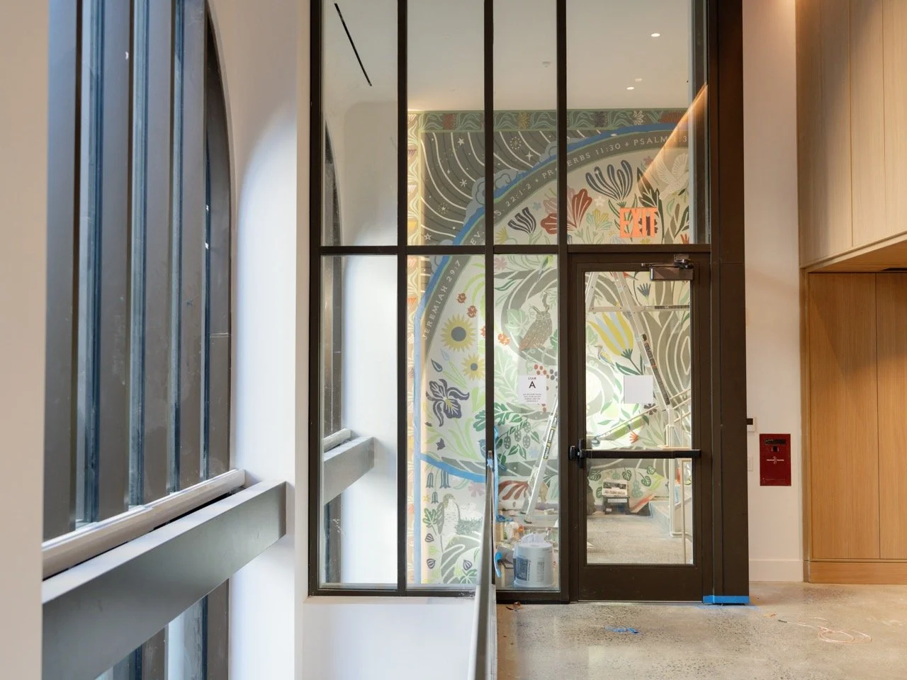 Interior view of a modern building with a black-framed glass door and window. Through the glass, a colorful mural with floral and abstract designs is visible. A ladder and painting supplies are in front of the mural, indicating ongoing artwork.