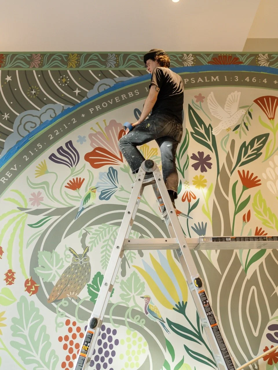 A person standing on a ladder painting a colorful mural on a wall that features flowers, plants, birds, and an owl.