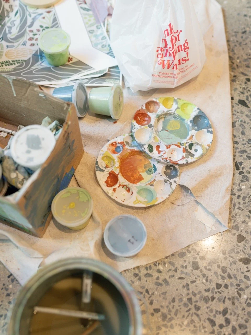 Paint palettes with multicolored paint, empty paint containers, a cardboard box with used paint tubes, a paper bag with red text, and paper with colorful designs on a table surface.