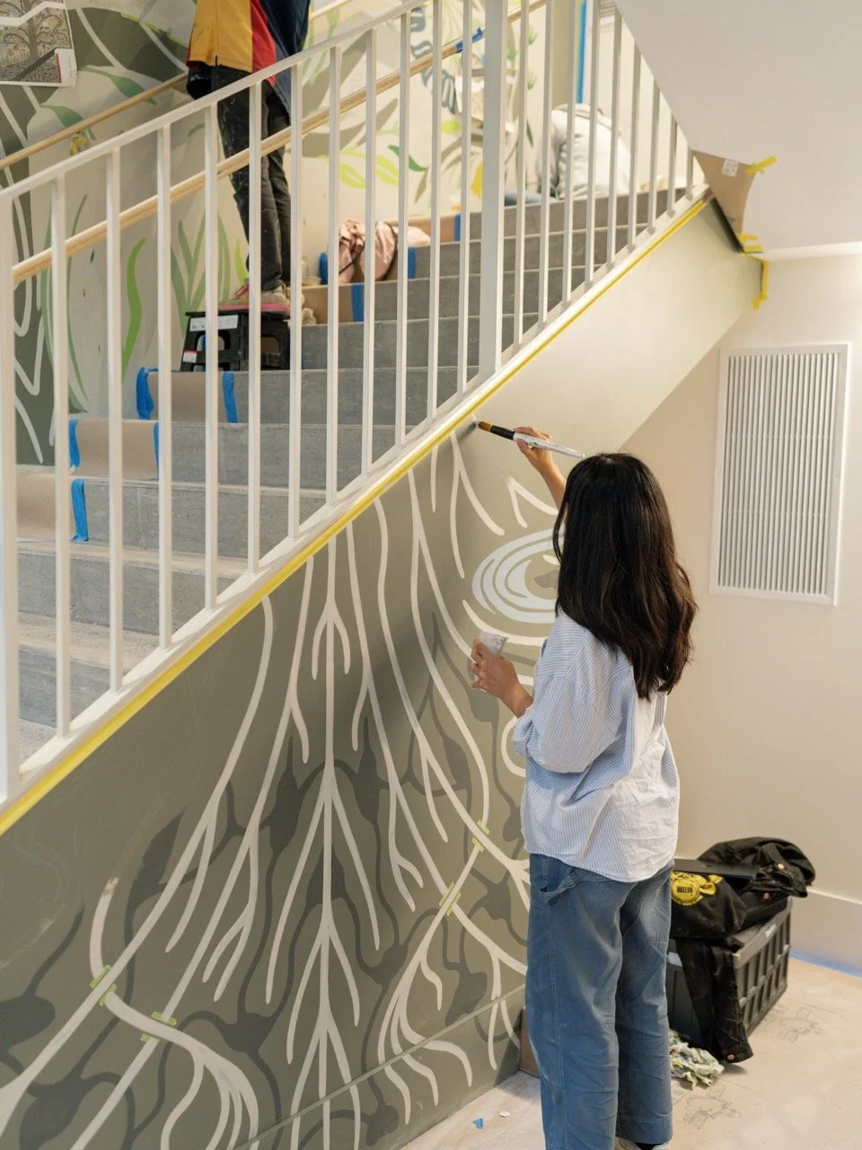 A woman is painting a mural of tree roots on the wall beneath a staircase, while a person on the stairs is working on the upper part of the wall or railing.