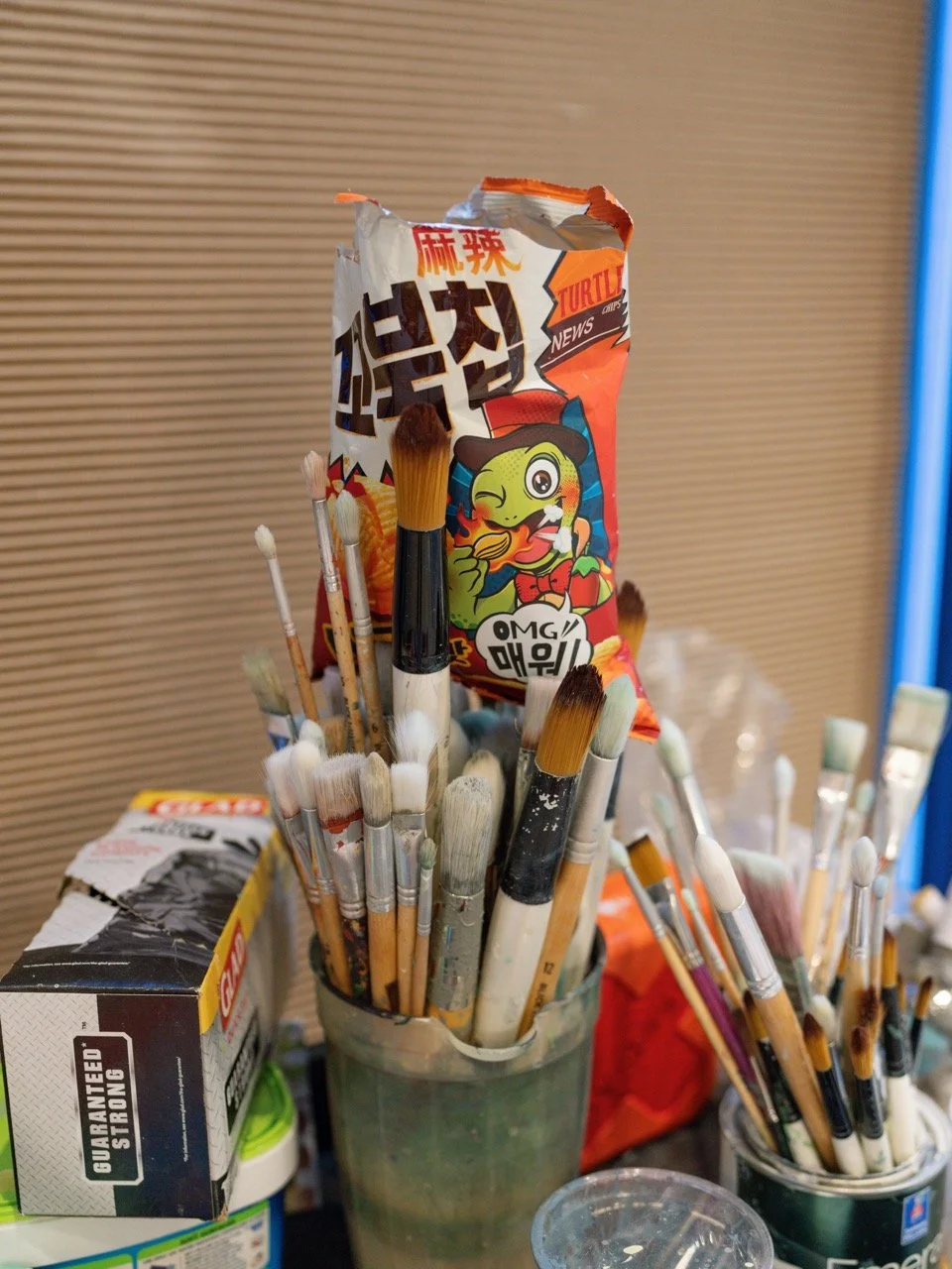 A jar filled with various paintbrushes and an open snack bag with a cartoon character on it, placed among other art supplies.