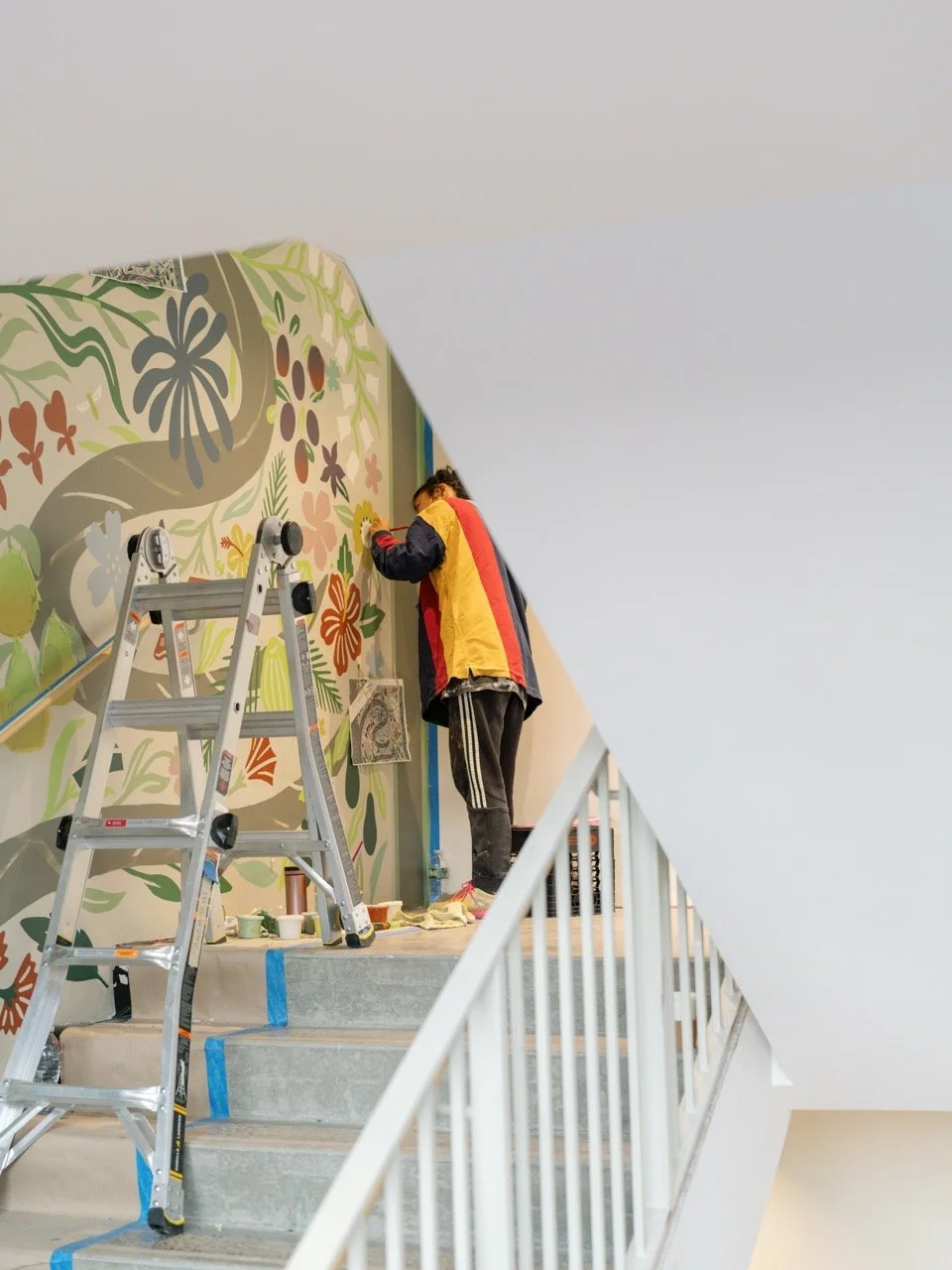Person painting a colorful mural of flowers and foliage on a wall, standing on steps next to a ladder.