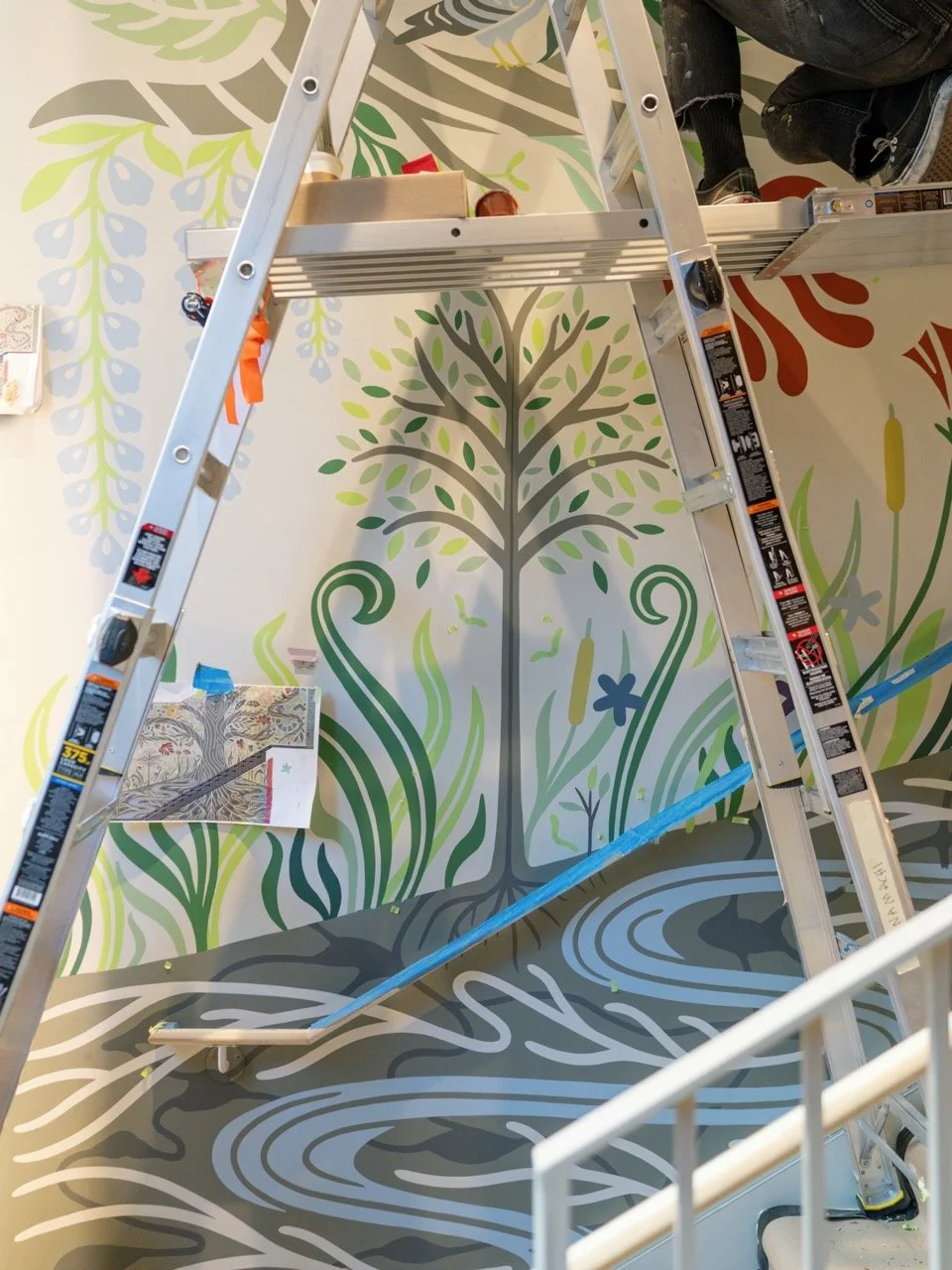 A ladder is placed over a colorful wall mural of a stylized tree with swirling roots, leaves, and small plants, with a person's legs visible in the top right corner.
