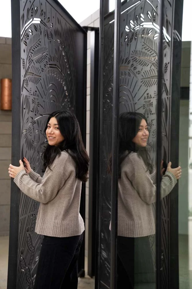 A woman with long dark hair smiling while holding onto black a decorative gate with intricate patterns, with her reflection visible in a mirror panel.