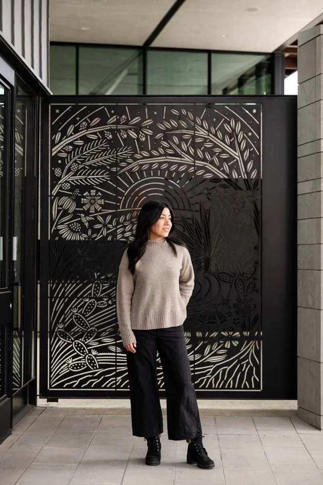 A woman standing in front of a decorative black metal entry gate with laser cut floral and botanical patterns attached to a modern building.
