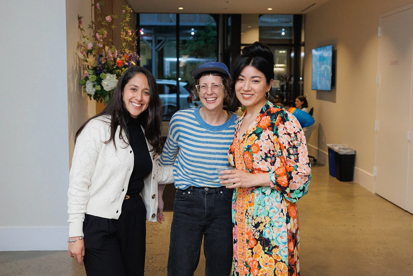 Three women smiling and posing together indoors, with a large flower arrangement and other guests in the background.