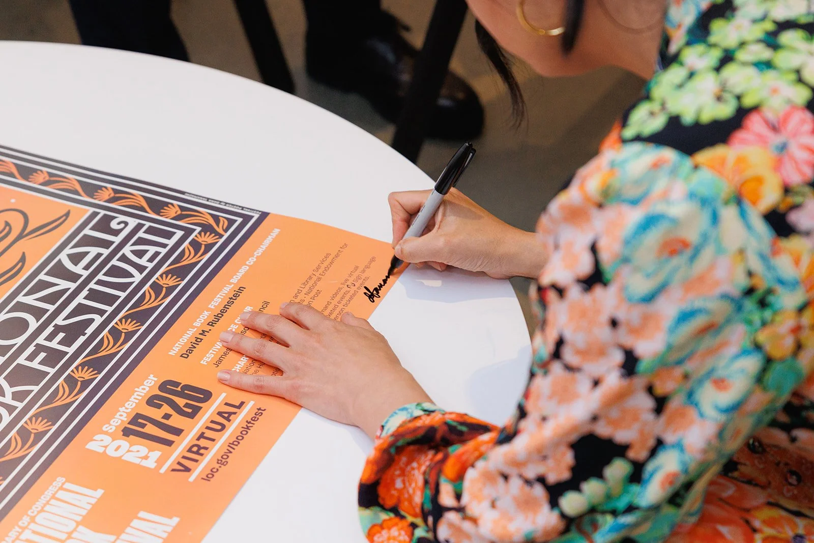 A woman in a colorful floral shirt is signing a poster for an event at a white round table.