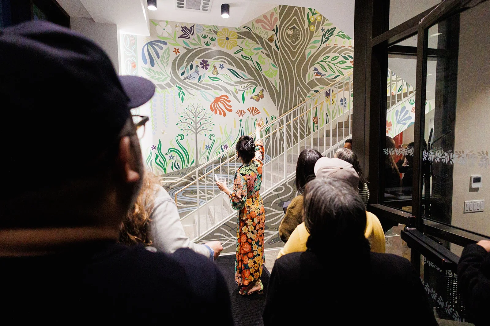 A group of people gathered indoors, listening to a woman in a floral dress who is speaking and gesturing towards a colorful wall mural with trees, animals, and butterflies.