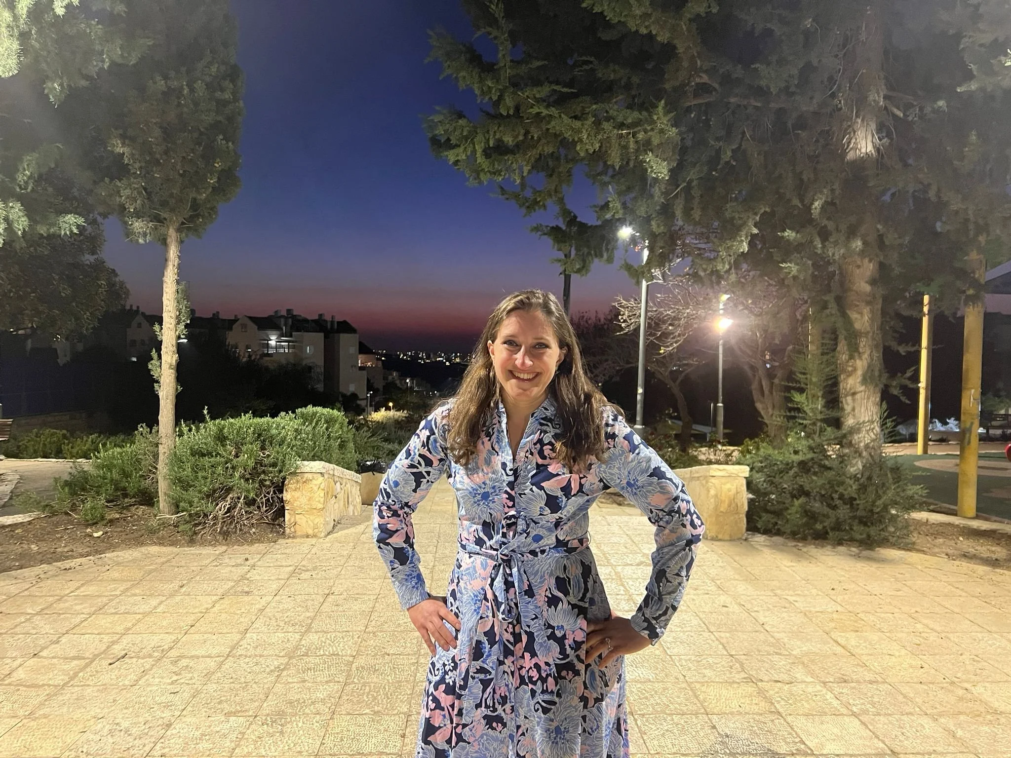 A woman in a long floral dress smiling outdoors at dusk or early evening. She has long brown hair and is standing on a paved area with trees and bushes behind her, with a cityscape visible in the distance.