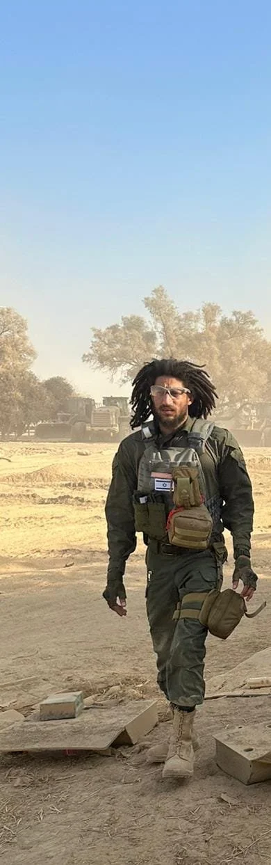 A man with dreadlocks wearing sunglasses and military gear walking through a desert area with trees and trucks in the background.
