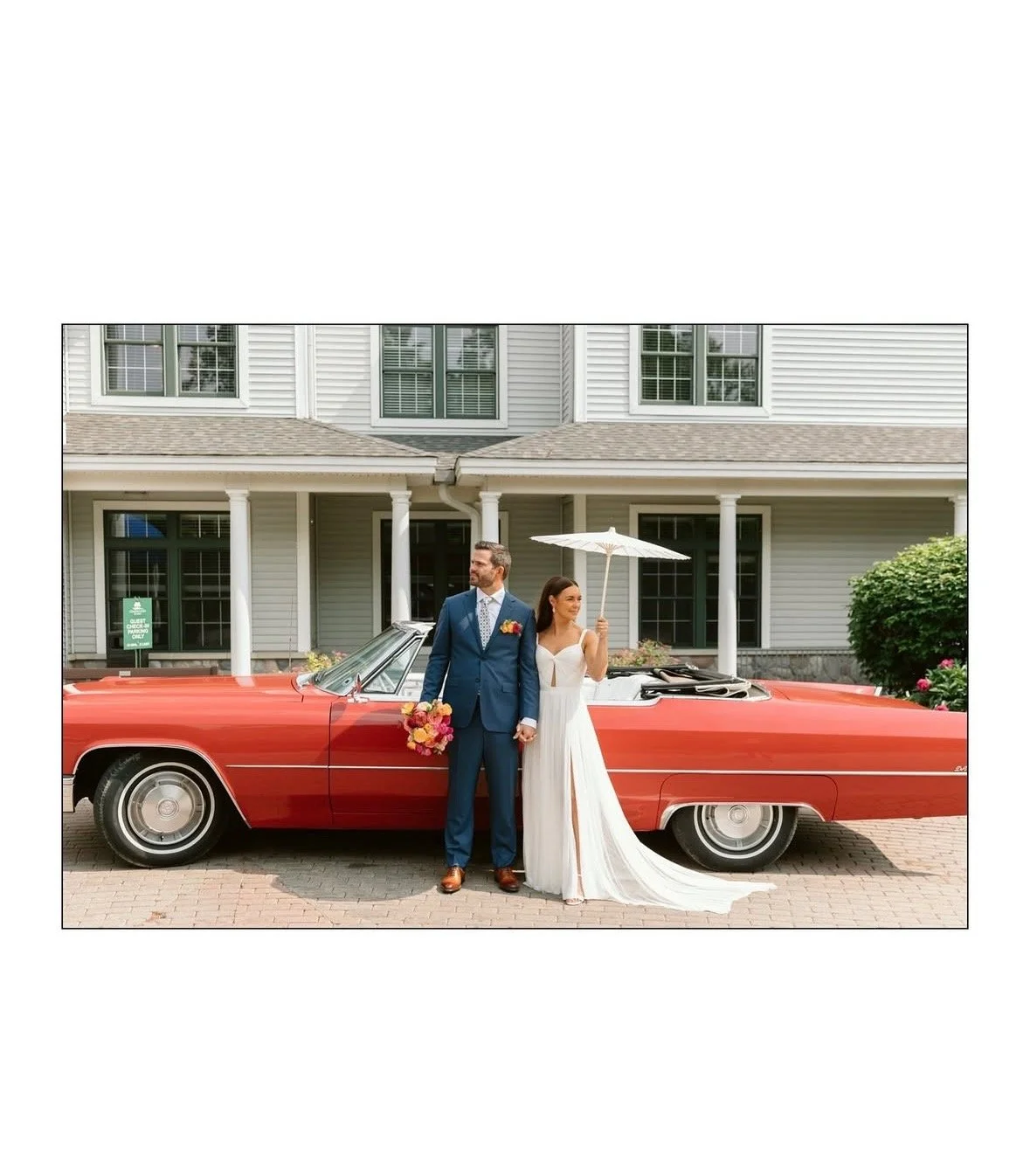 A bride and groom stand in front of a vintage red car on their wedding day, with the bride holding a white umbrella and the groom holding a bouquet.