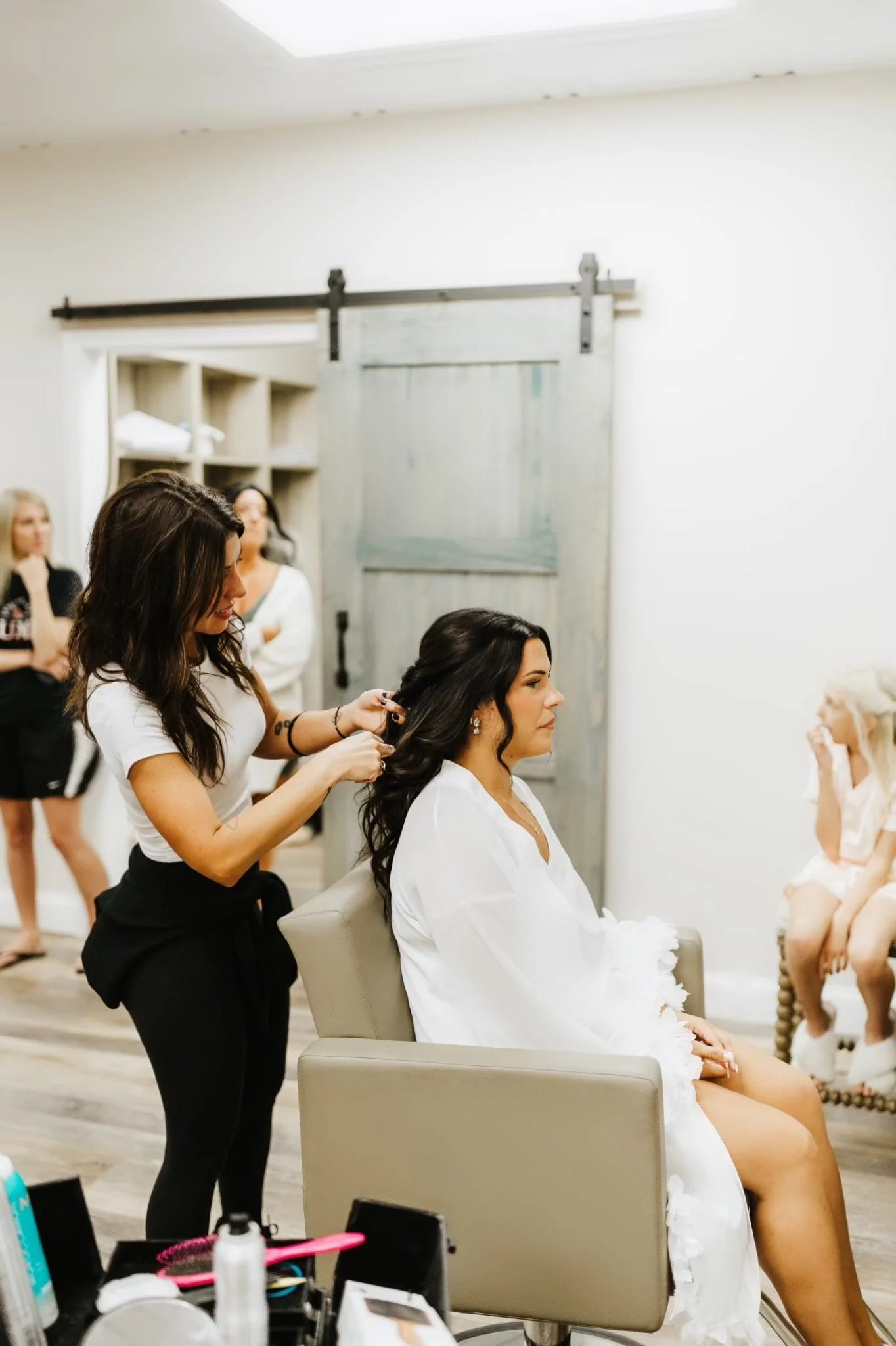 Women preparing for a wedding in a room, with a hairstylist working on a seated bride's long dark hair.
