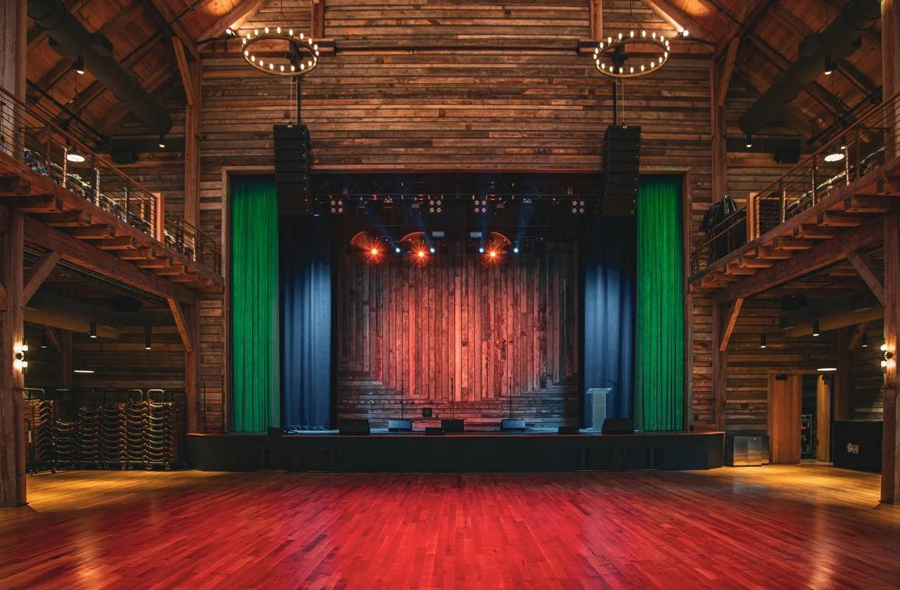 Empty stage in a rustic wooden venue with green and black curtains and colorful stage lights.