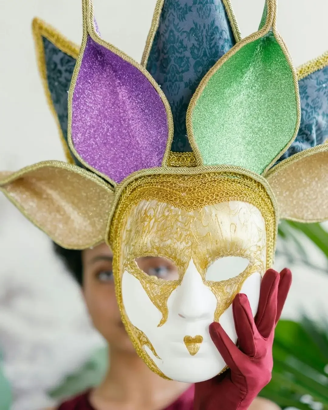 Persona sosteniendo una máscara de carnaval adornada con pétalos decorativos de colores brillante y borde dorado.
