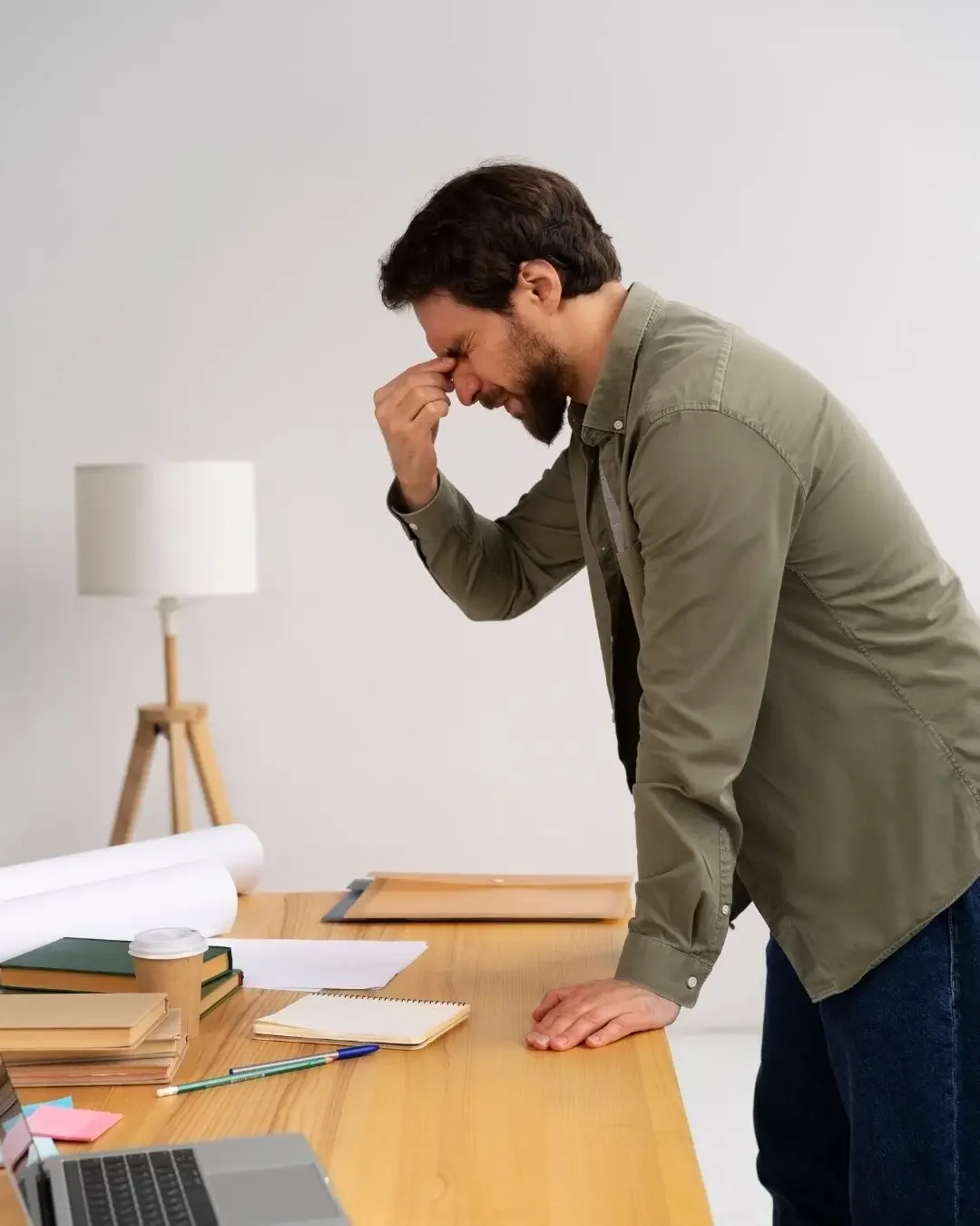 Hombre con expresión de dolor o incomodidad, sosteniendo la frente con la mano, en una oficina o espacio de trabajo con mesa de madera, libros, cuadernos, taza de café y laptop.