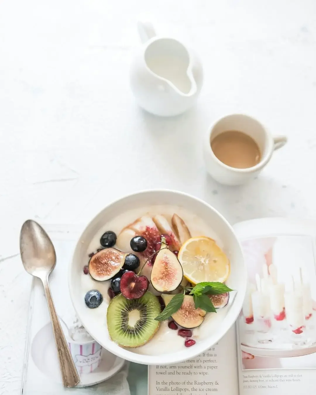 Cuencos de frutas con fresa, kiwi, uvas, y limón, además de una taza con café, un recipiente con leche y una cuchara sobre una mesa blanca.