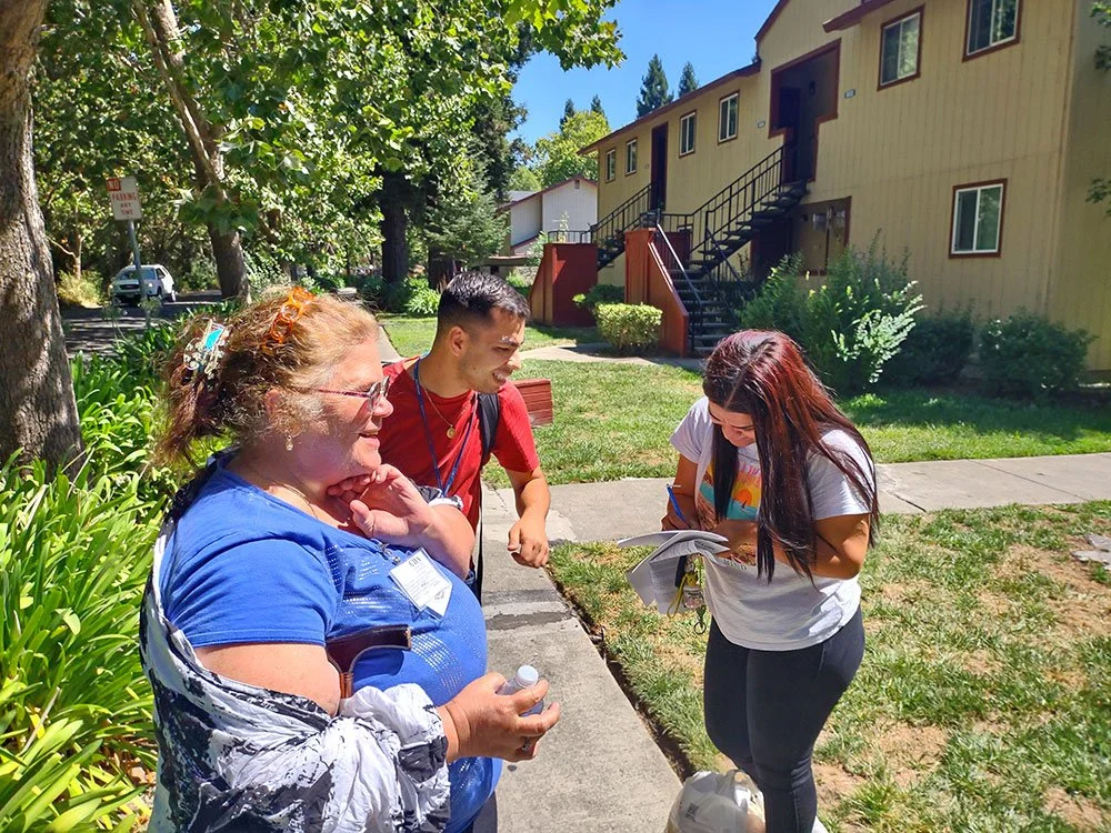 A smiling white female in a blue shirt and a smiling Latino male in a red shirt sign up a woman on the sidewalk near an apartment building.