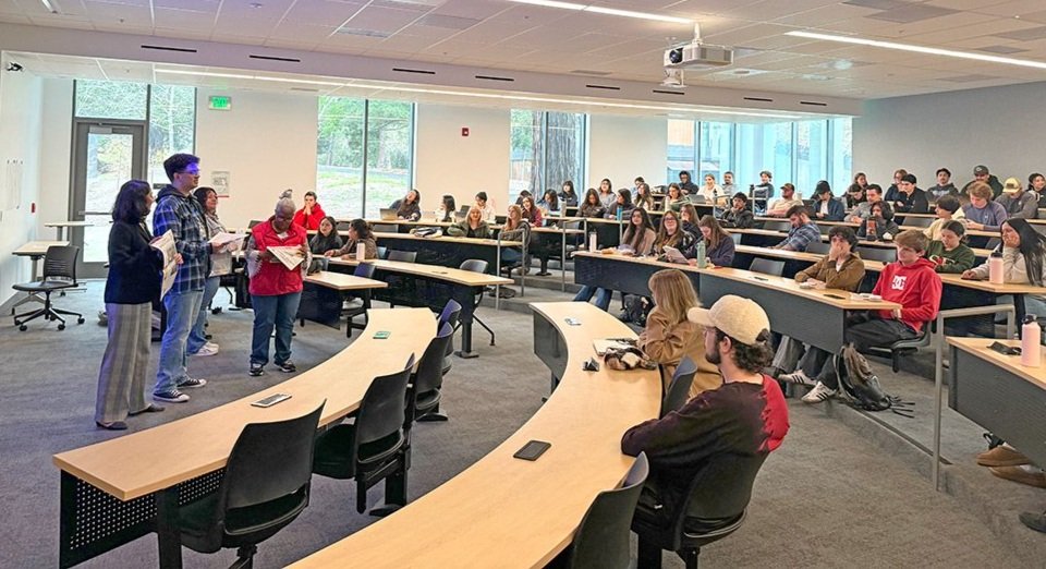 Four people speak to a university classroom of around 80 students.