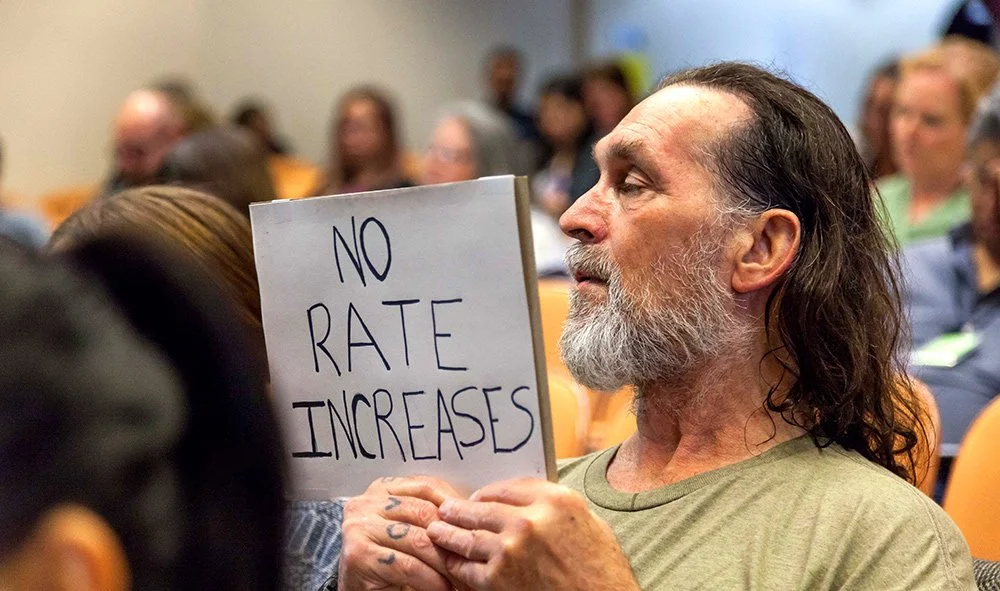 A man in a hearing room holding up a sign reading "NO RATE INCREASES"