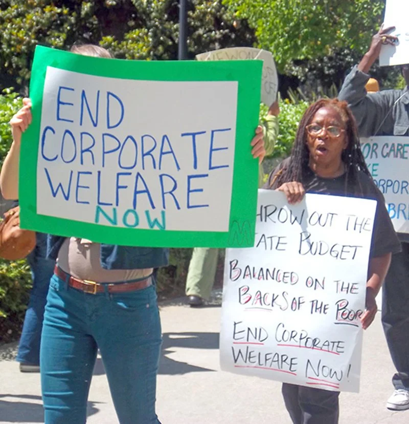 Multiple people on a picket line. One sign says "END CORPORATE WELFARE NOW"