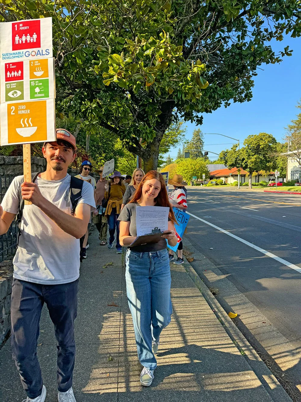 A young white male with a picket sign showign 4 of the 17 sustainable development goals and white female writing on a clipboard on a march on a sidewalk. Other people with signs are in the background.