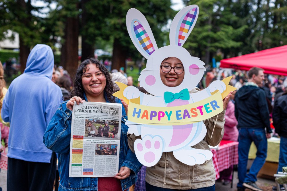 Two young Latina volunteers pose for a photo. One is holding a newspaper and the other is holding an Easter bunny with her face in place of the bunny's face.