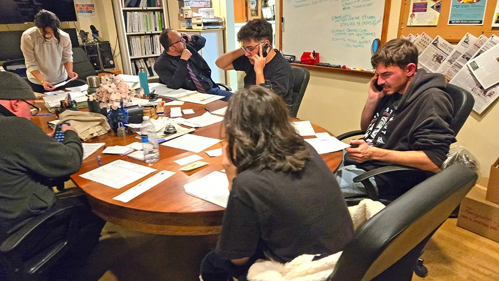 Four young people using telephones, around a table with papers and water bottles on it. Others are partially visible around the table as well.