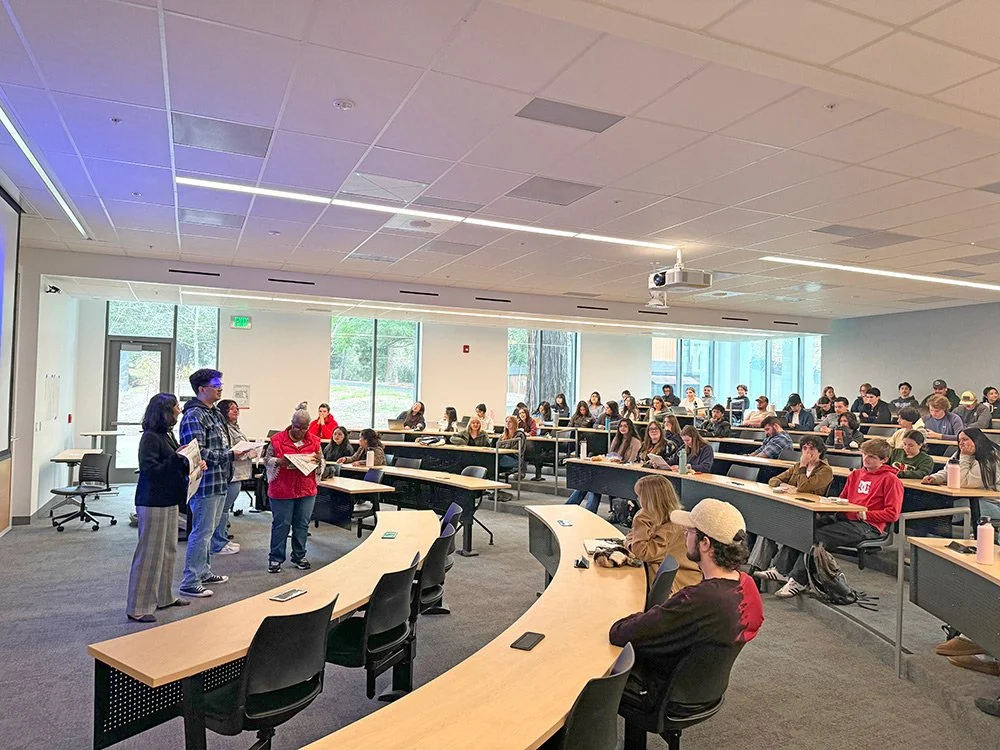 Four people speak to a university classroom of around 80 students.