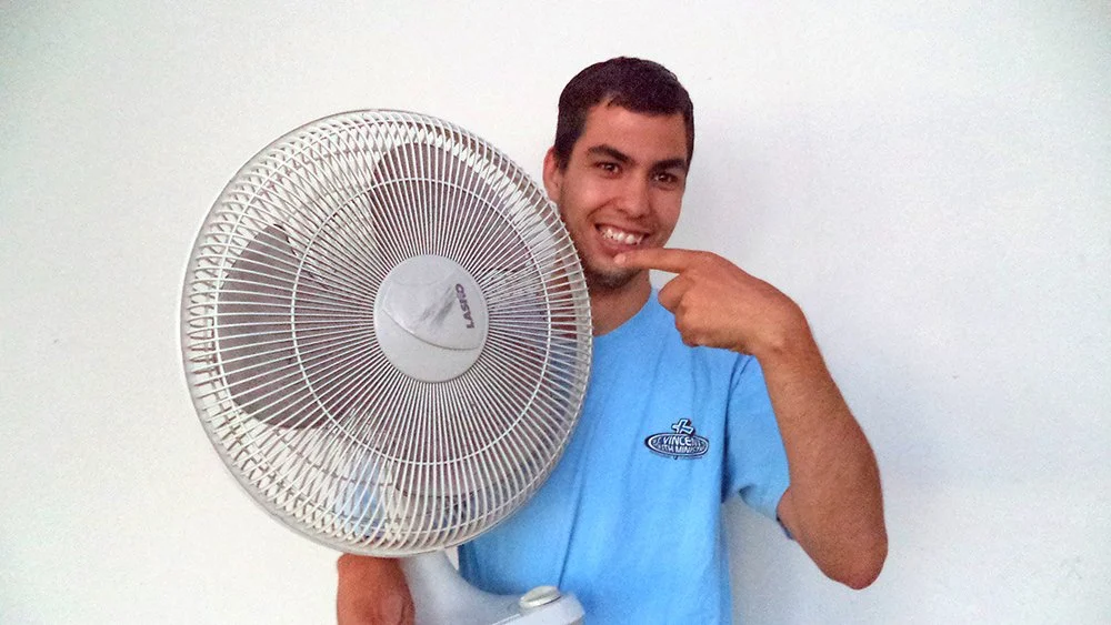 A smiling young Latino holding a table top fan in one arm and pointing to it with his other hand.