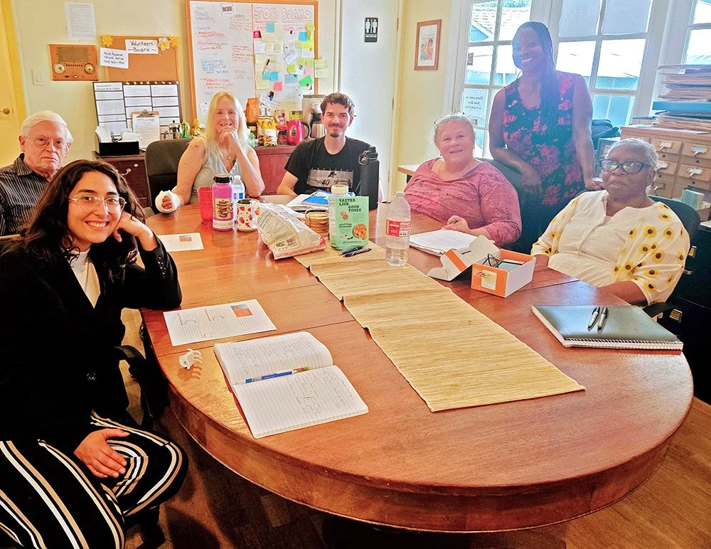 Four smiling people, three female and one male, sitting at or near a large wooden table.