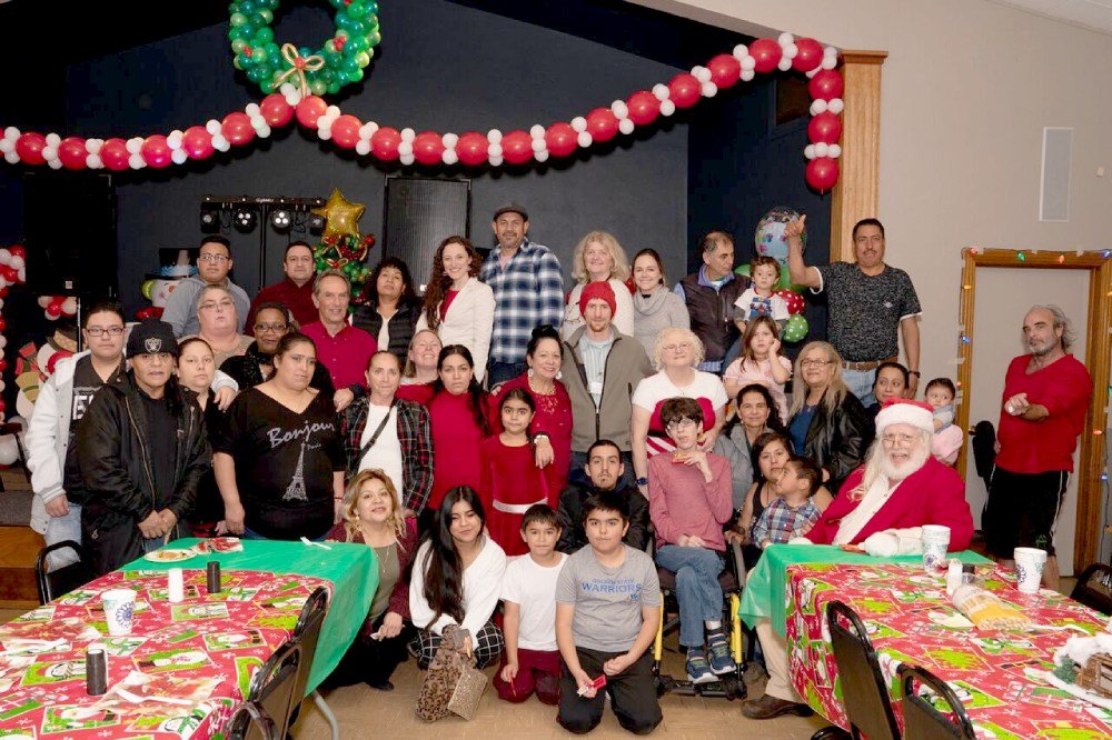 A group of about 40 mixed men, women and children at a holiday-themed event with decorated tables in front of them and a decorated stage behind them.