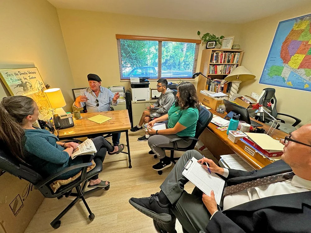 A group of five people around a desk work together. Some have clipboards and are taking notes. The sun is shining outside the window.
