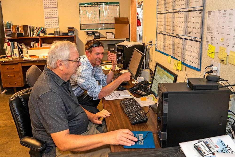 Two males work together at a desktop computer in an office.