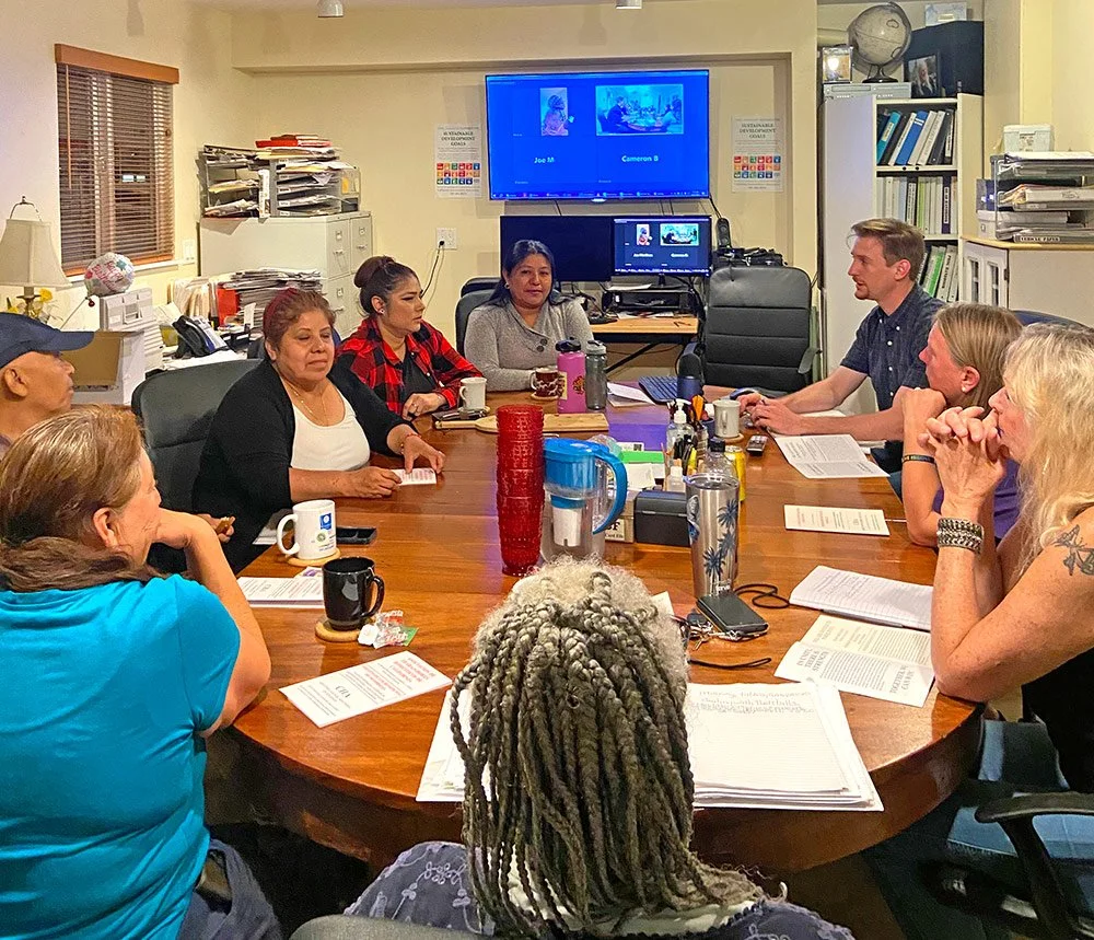 A group of mostly women at a large wooden table with a screen in the background showing a virtual conference.