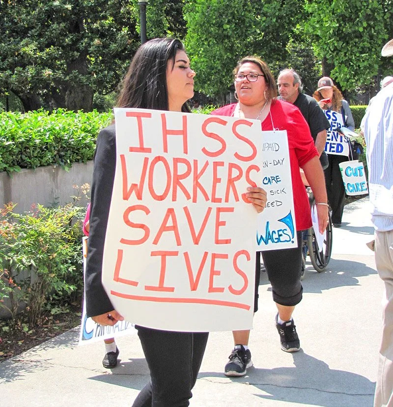 A young women on a picket line with a sign that says IHSS WORKERS SAVE LIVES in all caps and big letters.