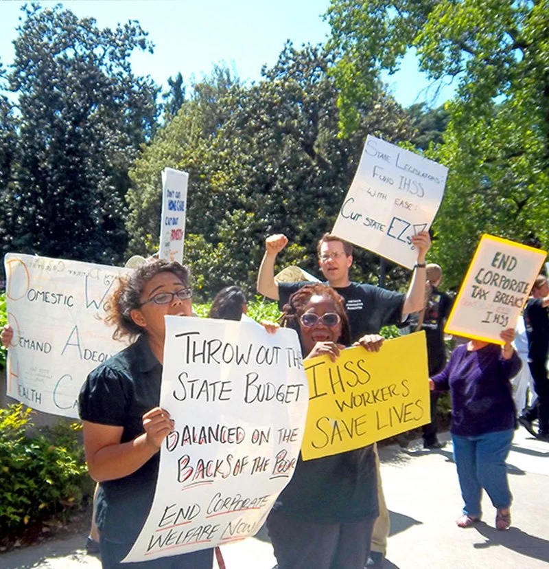 Multiple people with signs chanting on a picket line. The signs are against cuts to IHSS and state how the workers save lives.