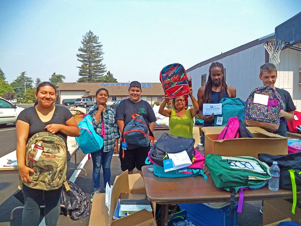 A smiling group of young mixed-race people holding up new backpacks outside.