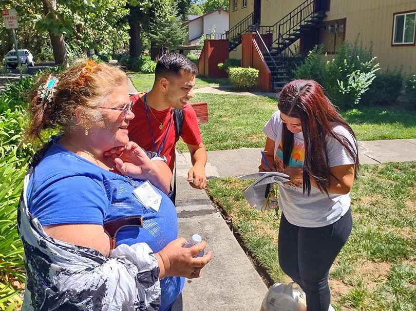 A white female in a blue shirt and Latino male in a red shirt sign up a woman on the sidewalk near an apartment building.