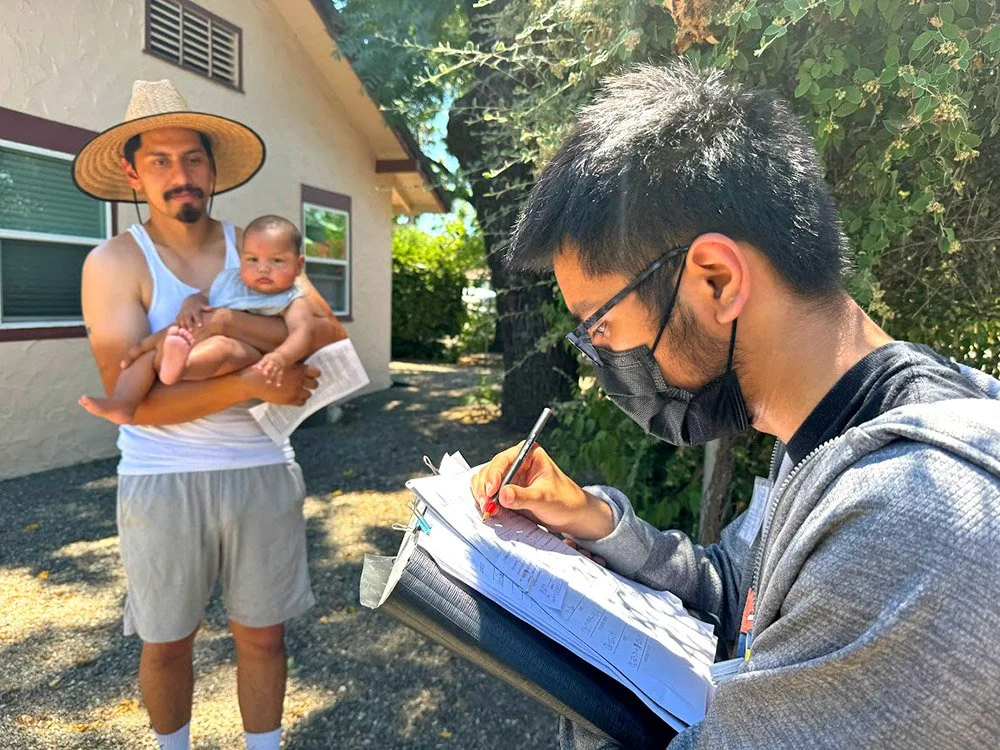 A young Vietnamese signs up a man holding a baby and a piece of paper in a hat in front of a house.